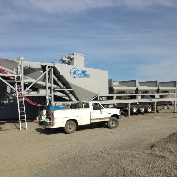 A white pickup truck is parked next to a large, industrial concrete batching plant with gravel piles nearby, under a clear blue sky. The equipment is labeled ACCUMIX LLC.