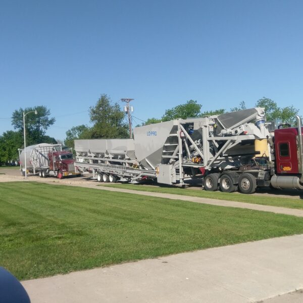 A large maroon semi-truck hauls oversized industrial equipment on a long trailer, parked on a street beside a grassy lawn, with buildings and trees in the background under a clear blue sky.