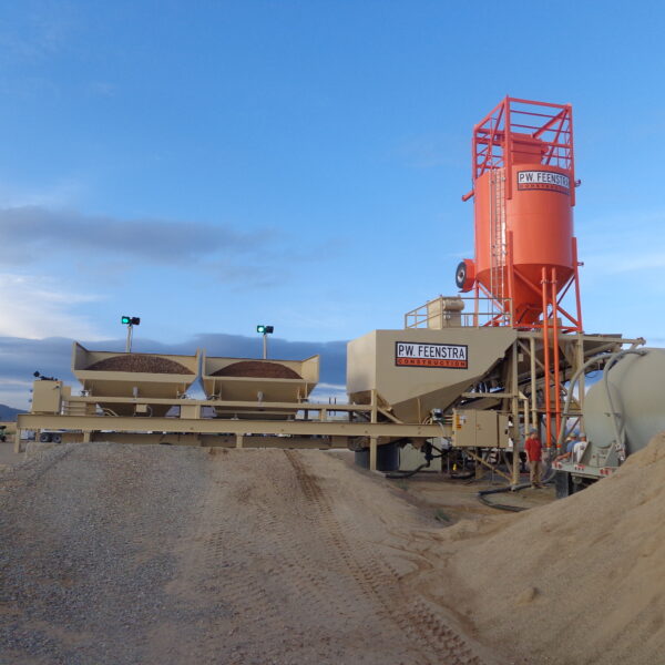 A concrete batching plant with gravel piles in the foreground, two aggregate hoppers, and a tall red cement silo labeled P.W. Feenstra set against a partly cloudy sky.
