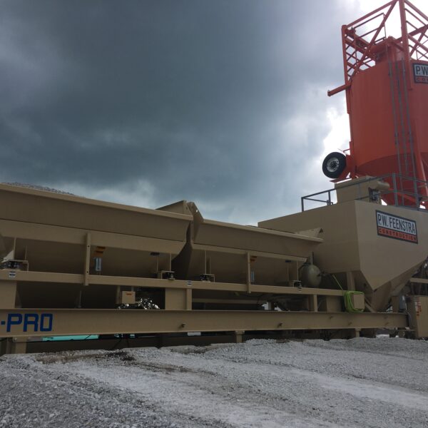 A concrete batching plant with tan and orange equipment, including storage silos and aggregate hoppers, stands beneath a dark, cloudy sky on a gravel-covered site.