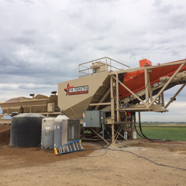 A large industrial machine labeled “D.L. Feenstra Construction” processes gravel or soil outdoors under a cloudy sky, with water tanks and hoses nearby and green fields in the background.