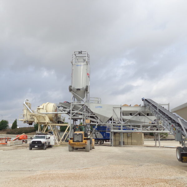 A concrete batching plant with large machinery, including silos, conveyors, trucks, and construction equipment, set on a gravel lot under a cloudy sky. Trees and a fence are visible in the background.