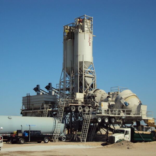 Large CON-E-CO wet batch central mix concrete batching plant with silos, conveyors, and machinery under a clear blue sky; trucks and equipment are parked around the structure on a gravel lot.