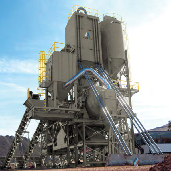 A large industrial asphalt mixing plant with tall silos, metal structures, and several pipes, standing outdoors against a clear sky with mountains in the background.