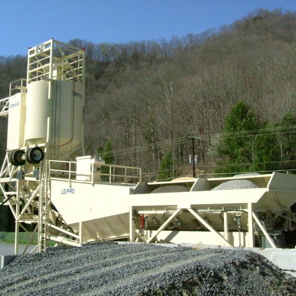 A concrete batching plant with silos, conveyors, and aggregate bins is set up outdoors near a wooded hillside under a clear blue sky. Piles of gravel are visible in the foreground.