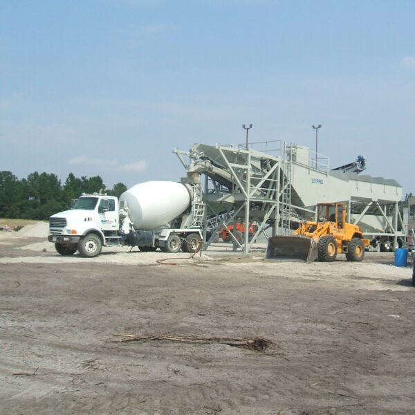 A concrete batch plant with a cement mixer truck, a yellow front loader, and piles of sand and gravel on a construction site under a clear sky. A car and a trailer are parked nearby.