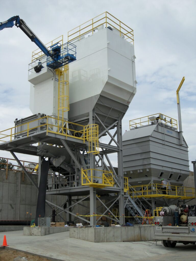 A large industrial transit mix concrete batch plant structure with white storage silos and yellow railings, supported by a gray steel framework. A blue crane and workers are on site, and a truck is parked nearby. The sky is cloudy.
