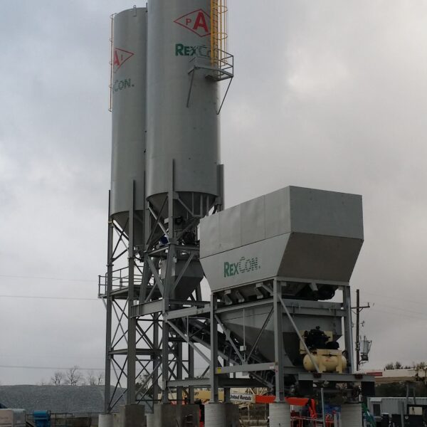 A large industrial concrete batching plant with two tall silos and a rectangular mixing unit, set on a construction site with dirt ground and cloudy skies.