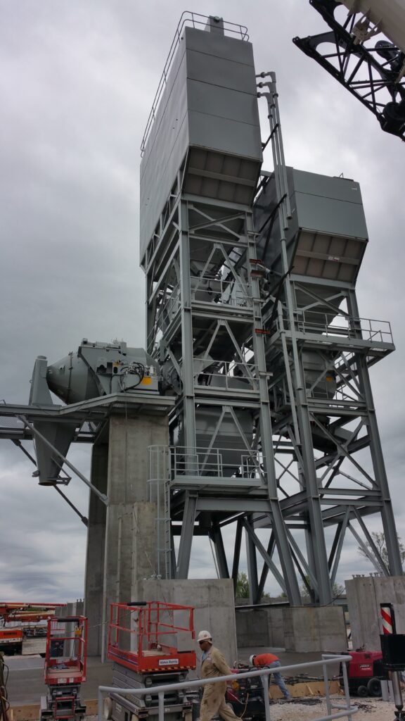 A tall industrial structure made of metal beams and large containers stands on a concrete base at a construction site; workers in hard hats and lifts are visible in the foreground under a cloudy sky.