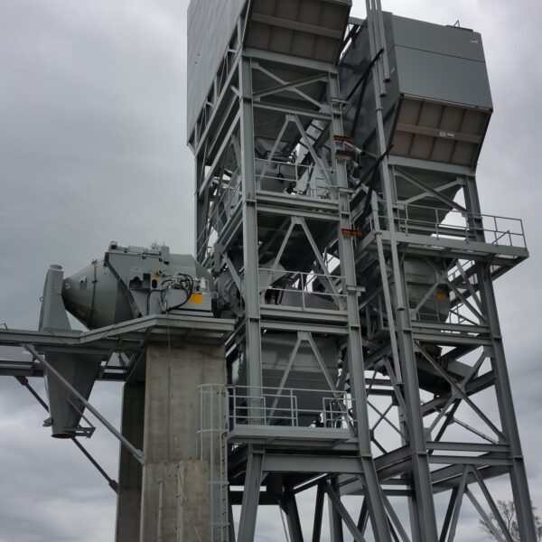 A tall industrial structure made of metal beams and large containers stands on a concrete base at a construction site; workers in hard hats and lifts are visible in the foreground under a cloudy sky.