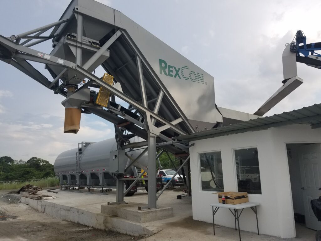 A large industrial machine labeled RexCon is positioned above a concrete platform near a small white building, with a truck and grassy area visible in the background.