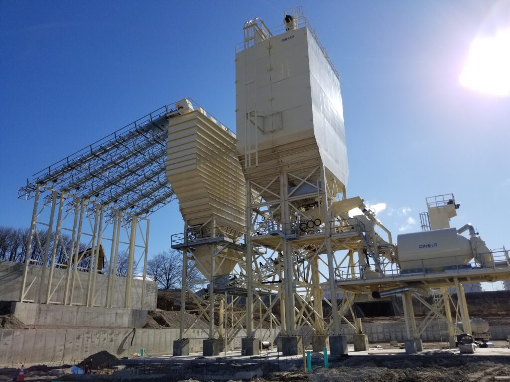 Large industrial stationary central mix concrete batching plant with cement bins and aggregate bins, horizontal reversing mixer, conveyor belts, and metal structures, set on a construction site under a clear blue sky.