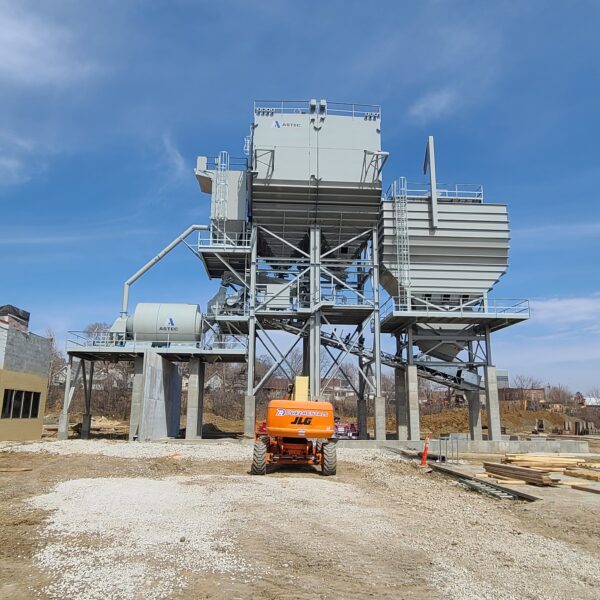 A large industrial structure with metal platforms and storage containers stands on concrete supports. A bright orange construction vehicle is parked in front, with a clear blue sky and trees in the background.