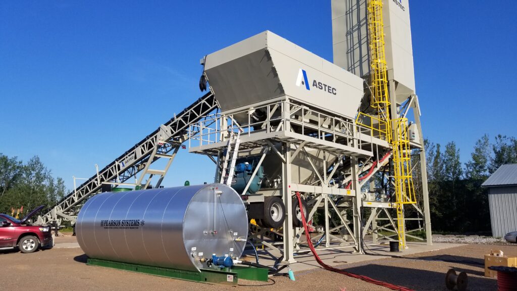 A large industrial Astec concrete batching plant with a metal tank labeled Pearson Water Heating Systems in front, conveyor belts, and a tall vertical structure, set outdoors on a sunny day.