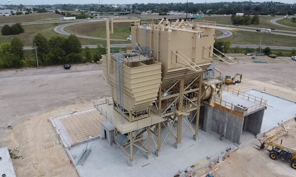 A large tan industrial asphalt plant with multiple storage bins and conveyors stands on a concrete platform at a construction site, surrounded by dirt, grass, roads, and vehicles in the background.