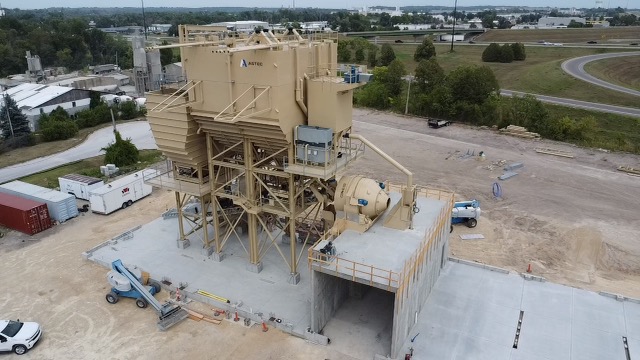Aerial view of a large ASTEC central mix concrete batching plant on a construction site, surrounded by vehicles, containers, greenery, and a nearby road.