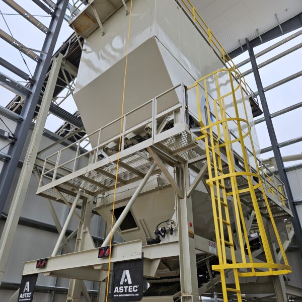 Large industrial ASTEC equipment stands inside a factory, featuring a white metal hopper, yellow ladders and railings, with steel beams and a high ceiling in the background.