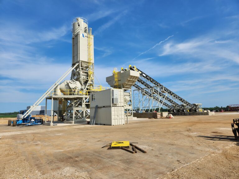 A large industrial Astec RexCon wet batch central mix concrete batching plant with silos, conveyors, and machinery stands on a wide, cleared construction site under a bright blue sky with scattered clouds.