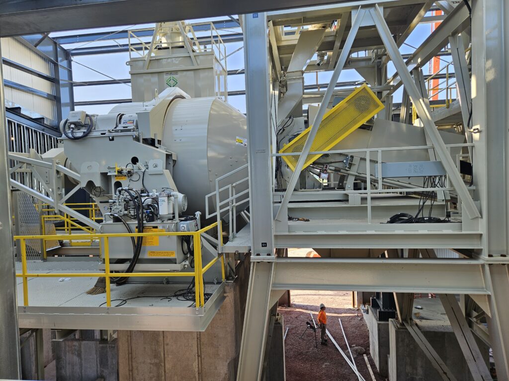 A large industrial central mix concrete plant with metal steel building framework and yellow railings inside a factory. A worker in an orange vest and hard hat stands below on a gravel area, emphasizing the scale of the equipment.