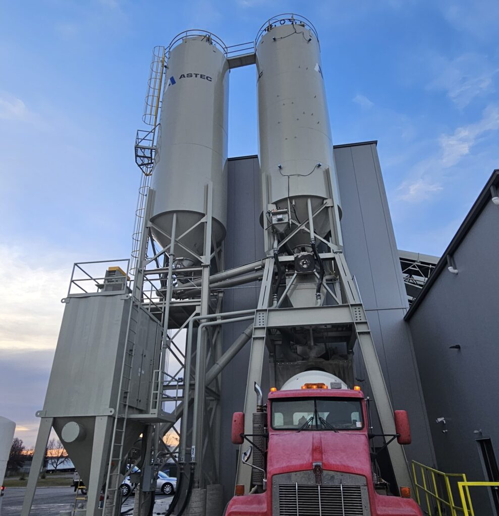 A red ready mix truck is parked in front of two tall industrial silos and a gray building at sunset. The sky is partly cloudy with a blue hue. The silos have the word ASTEC printed on one of them.