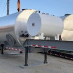 Two large, silver industrial storage tanks on trailers are parked outdoors near a utility pole and a building under a clear blue sky. A white truck is partially visible on the right.