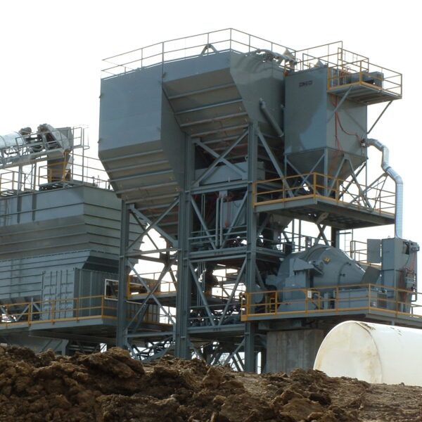 Large industrial asphalt plant with metal silos, conveyors, and support structures, surrounded by dirt and a white tank in the foreground, set against a bright sky.