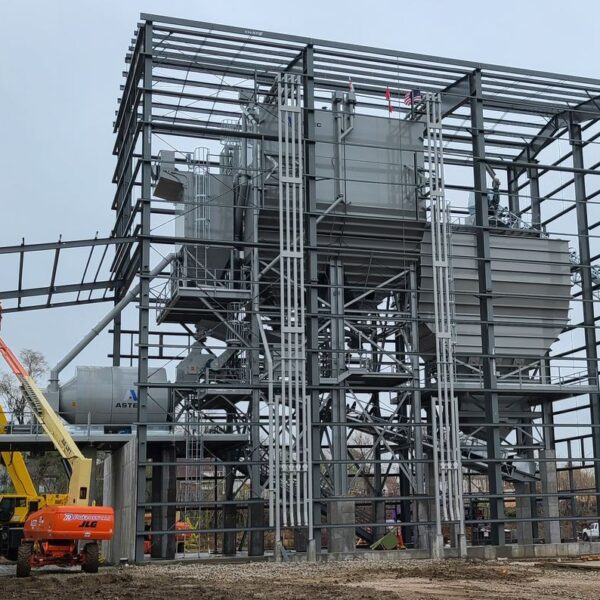 A large steel industrial structure under construction, with cranes and aerial lifts in use. The frame is partially complete, and a conveyor belt extends from the right side. Workers and construction equipment are visible on site.