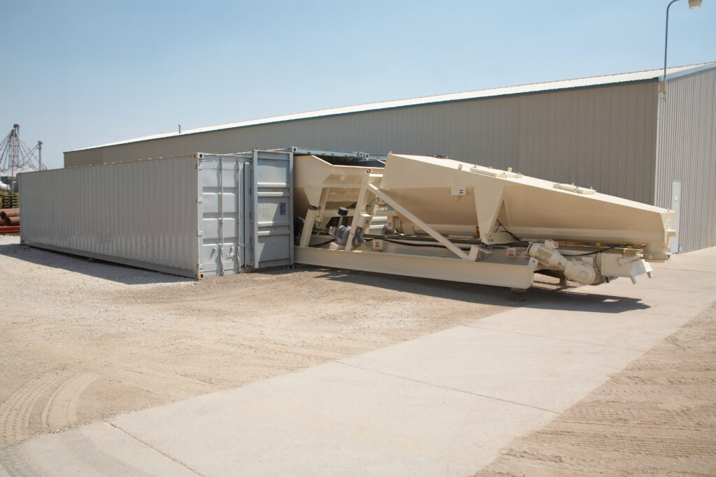 Large beige industrial equipment is partially inside a gray shipping container next to a metal warehouse on a clear day. The surrounding area is unpaved with some dirt and gravel.