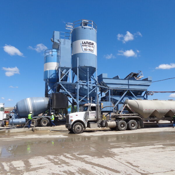 Trucks and workers are at a concrete batch plant with large blue silos and equipment under a clear blue sky. The ground is wet, and the facility is labeled LARSON Ready Mix.