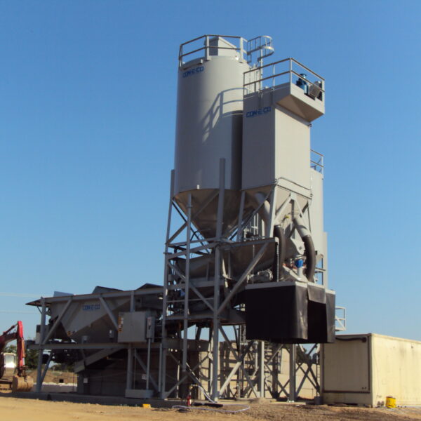 An industrial concrete batching plant with large silos, metal structures, and conveyor belts stands on a dirt lot under a clear blue sky. A small excavator is visible to the left, and storage containers are on the right.