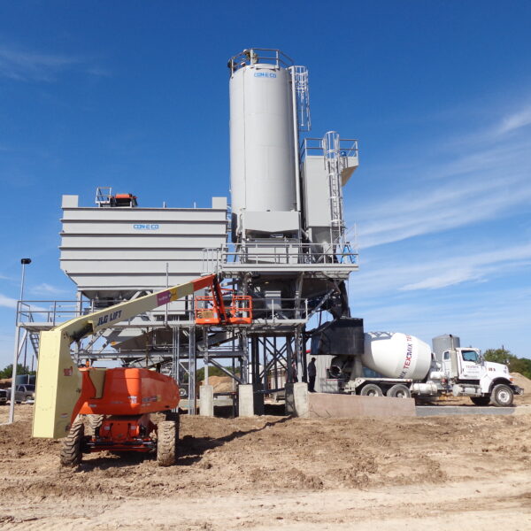 A concrete batching plant with tall silos stands on a construction site under a blue sky. A concrete mixer truck and an orange construction lift are nearby, surrounded by dirt and construction materials.
