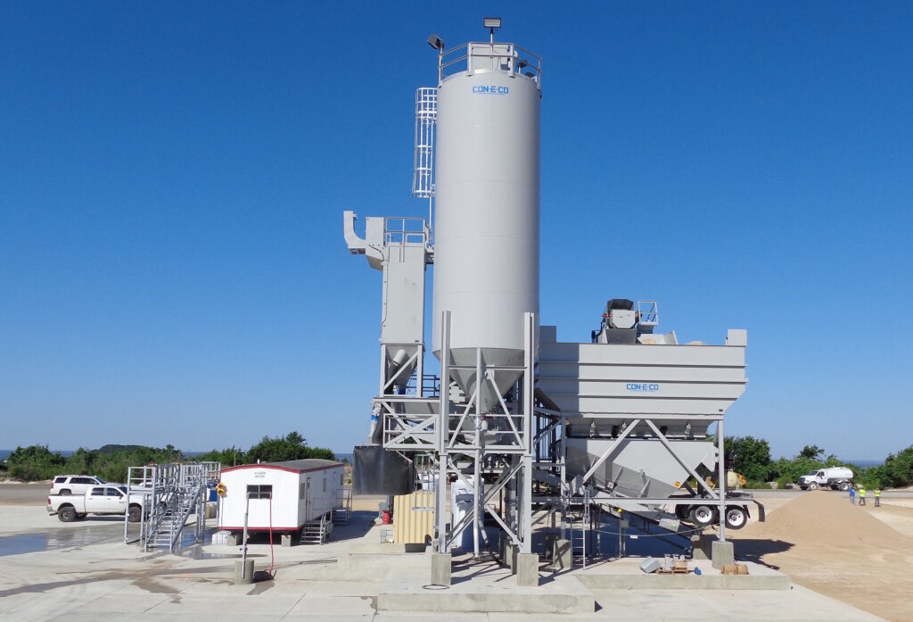 A large gray CONECO stationary dry batch concrete batching plant stands on a paved lot under a clear blue sky, with a white truck, trailer, and piles of sand nearby. Trees and greenery are visible in the background.