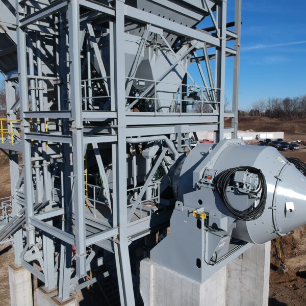 A large industrial machine with gray metal framework and a cylindrical component is mounted on concrete supports at a construction site, with bare trees and vehicles in the background under a clear blue sky.