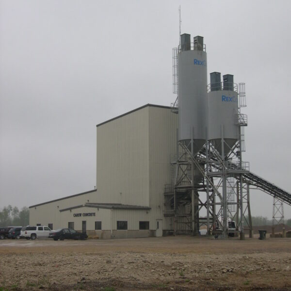 A concrete plant with tall silos labeled RexCon and a beige industrial building; several cars are parked outside on a cloudy, overcast day.