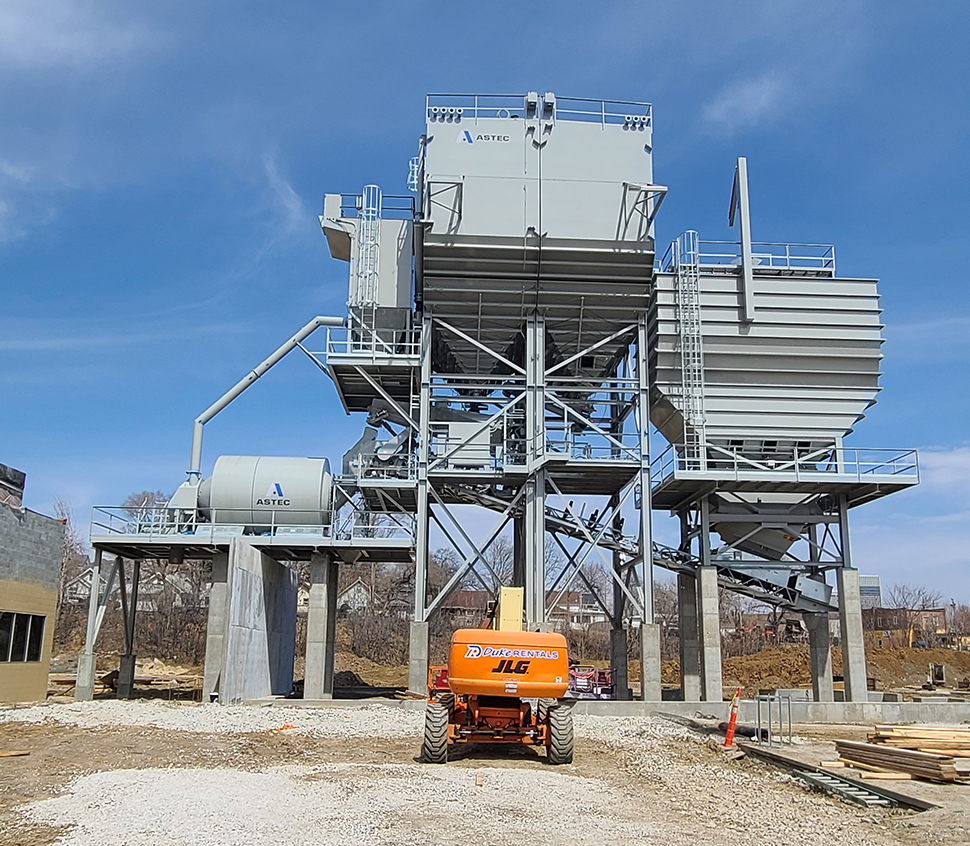 A large industrial central mix concrete batch plant with multiple cement bins and aggregate bins with batch belt conveyors stands on a construction site. An orange JLG lift is parked on gravel in front, with a clear blue sky and some trees in the background.