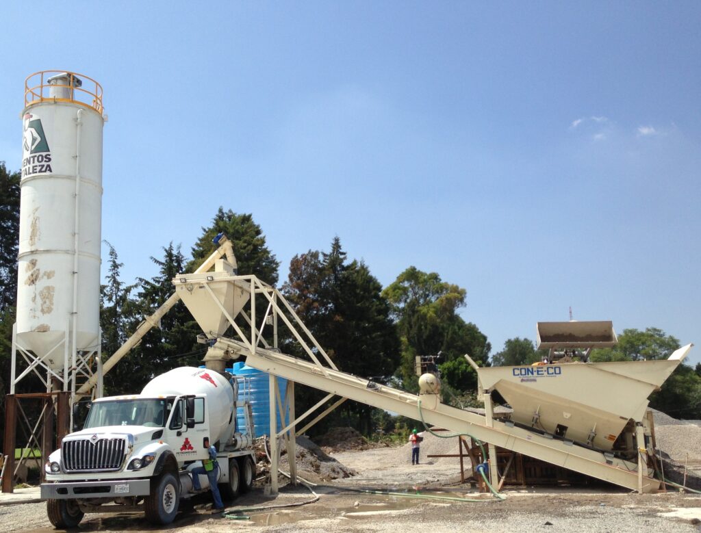 A cement mixer truck is parked at a concrete batching plant with silos, conveyors, and hoppers on a gravel lot. Trees and a clear blue sky are in the background.