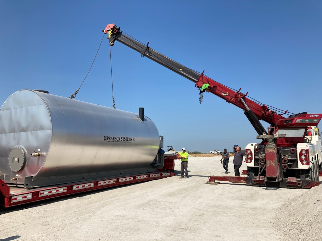A red crane lifts a large silver industrial tank labeled Pearson Water Heating Systems onto a flatbed trailer, while three workers in safety gear supervise on a construction site under a clear blue sky.