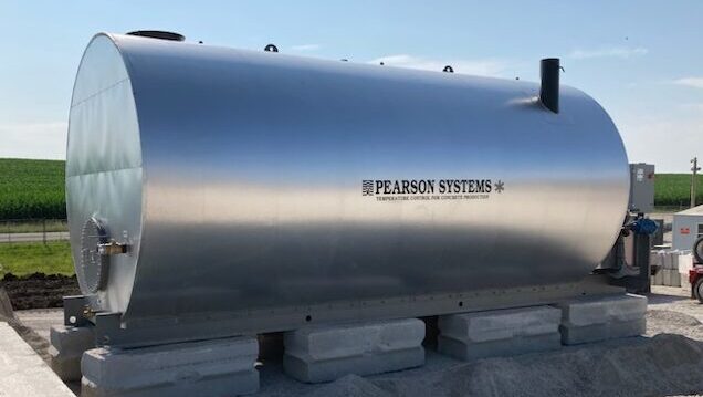 A large, cylindrical steel insulated water storage tank labeled Pearson Systems sits outdoors on concrete blocks, surrounded by gravel and construction equipment, with green fields visible in the background under a blue sky.