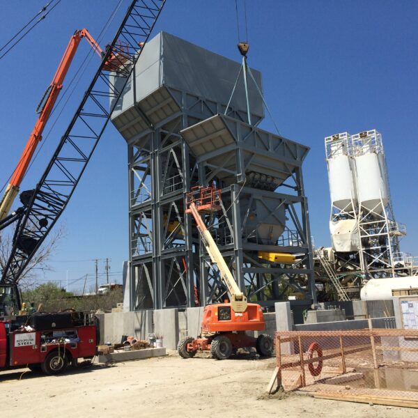 A construction site with cranes, trucks, and heavy machinery assembling a large industrial structure under a clear blue sky. Workers and building materials are visible around the site.