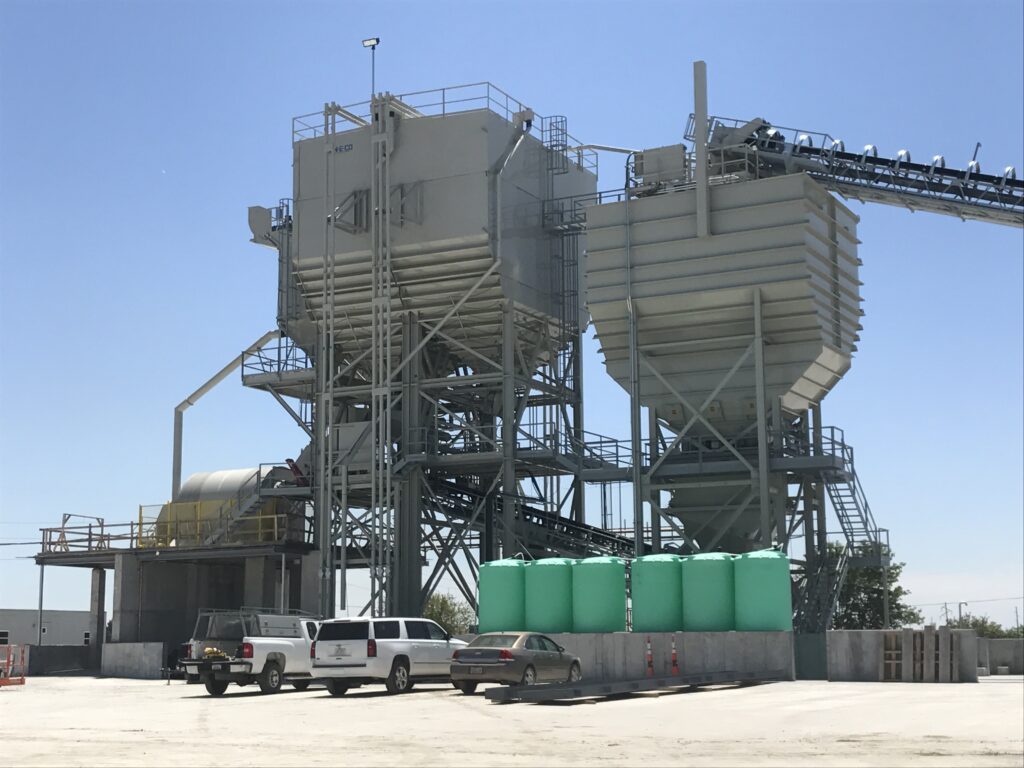 Two large industrial silos with supporting metal structures, conveyor belts, and green storage tanks stand in a concrete lot under a clear blue sky. Several parked vehicles are visible in the foreground.