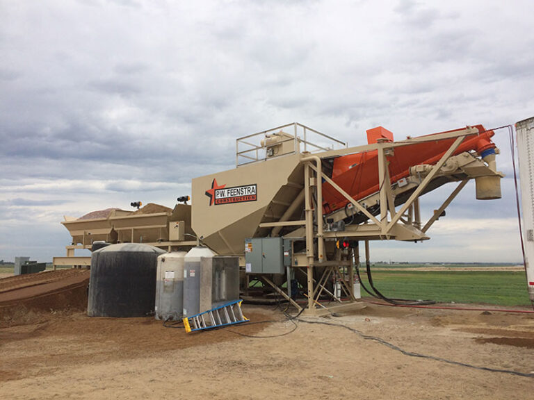 A large industrial cement gravel and sand mixing portable concrete batch plant for mixing concrete with ready mix trucks sits on dirt under a cloudy sky, surrounded by open fields.