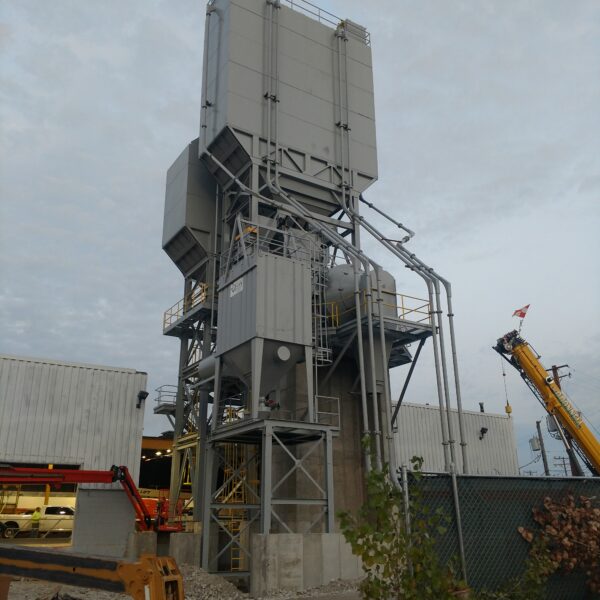 A tall, gray industrial structure with pipes and platforms stands outdoors near white buildings, construction equipment, and a green fence under a cloudy sky.