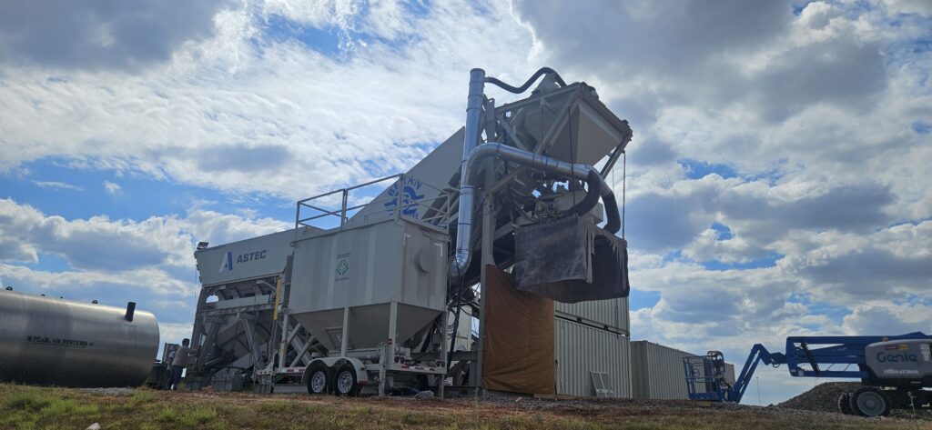 A large industrial C&W CP2100M central dust collector designed as a concrete batch plant stands outdoors on a construction site under a cloudy, blue sky. Machinery and tanks are visible nearby.
