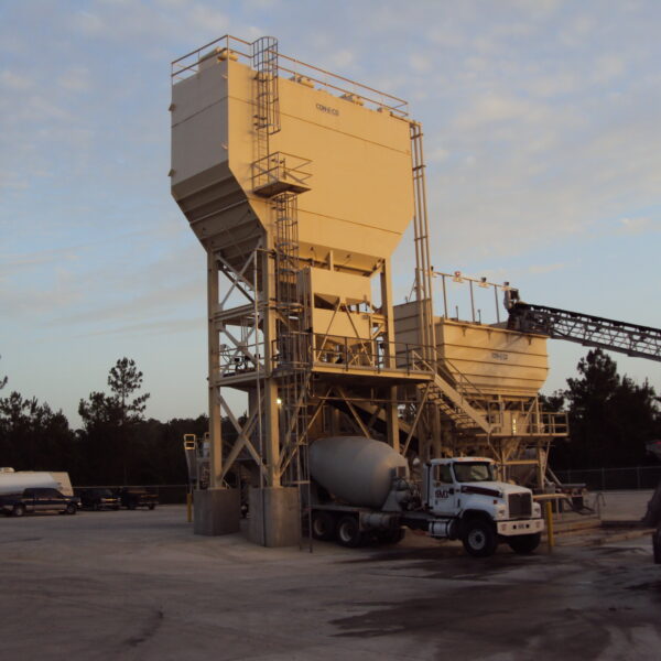 A concrete batching plant with large storage silos and a conveyor system, surrounded by cement mixer trucks and trailers, set against a backdrop of trees and a partly cloudy sky at sunset.
