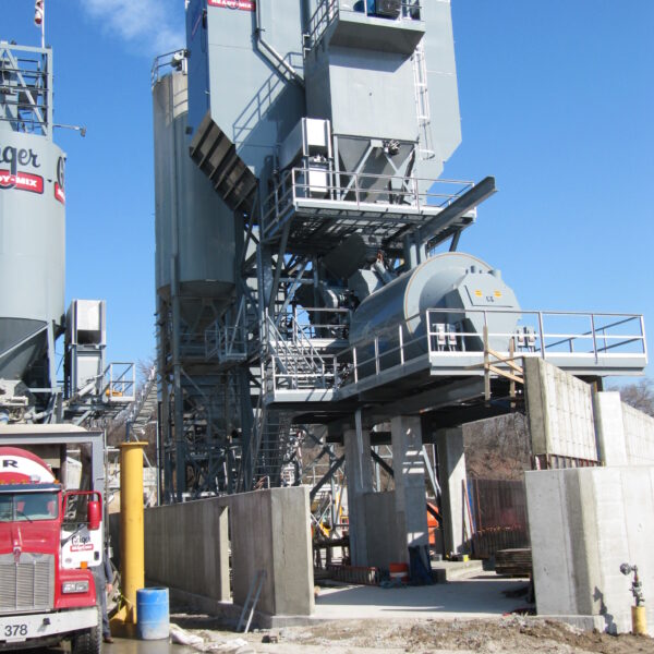 A large, gray concrete batching plant with multiple silos and metal structures under a clear blue sky, with trucks, barriers, and construction equipment in the foreground.
