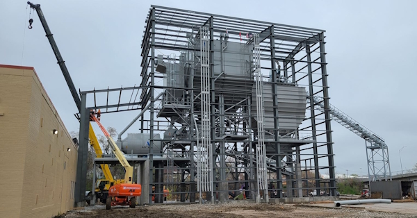 A large industrial steel structure under construction, with cranes and lifts on site. Two workers are elevated in a cherry picker, and a conveyor system extends from the structure. The sky is overcast.