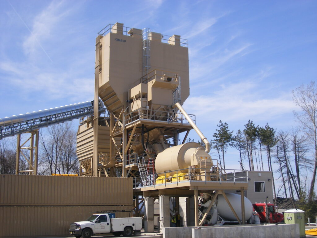 A large beige industrial asphalt mixing plant with silos, storage tanks, and elevated walkways, surrounded by vehicles and trees under a blue sky.