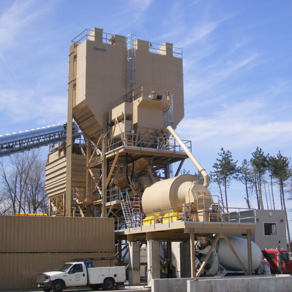 A large beige industrial asphalt mixing plant with silos, storage tanks, and elevated walkways, surrounded by vehicles and trees under a blue sky.