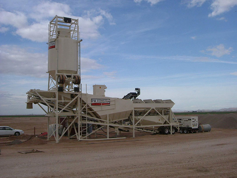Large portable concrete batch plant with silos and conveyors set up on a dirt lot, with a car parked nearby and open sky above. The equipment is white and appears to be used for mixing and loading concrete.