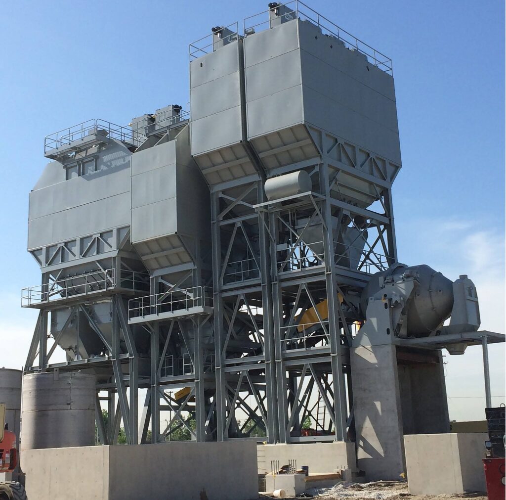 Large industrial machinery with multiple gray metal structures, tanks, pipes, and support beams, standing outdoors on a concrete base under a clear blue sky.